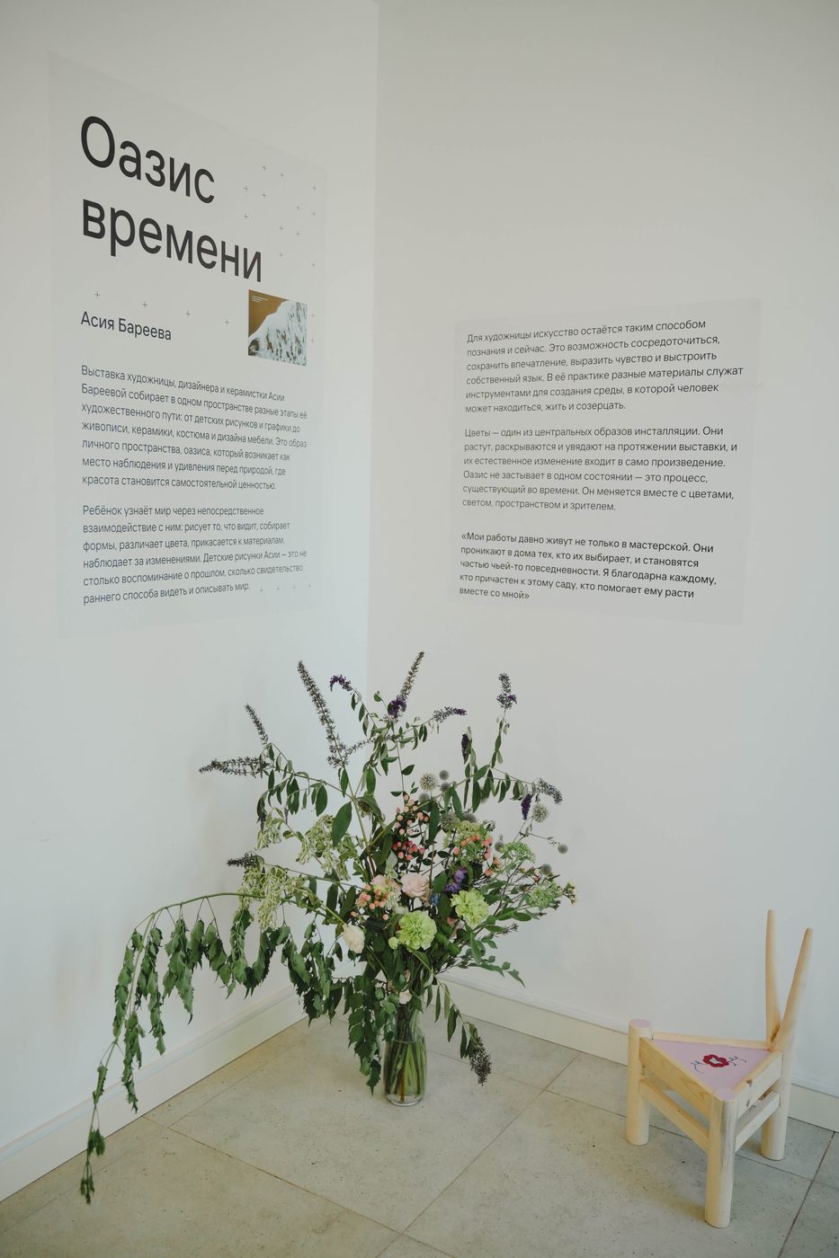 Экспликация выставки «Оазис времени» на стене, рядом на полу большой букет полевых цветов и детский стул
