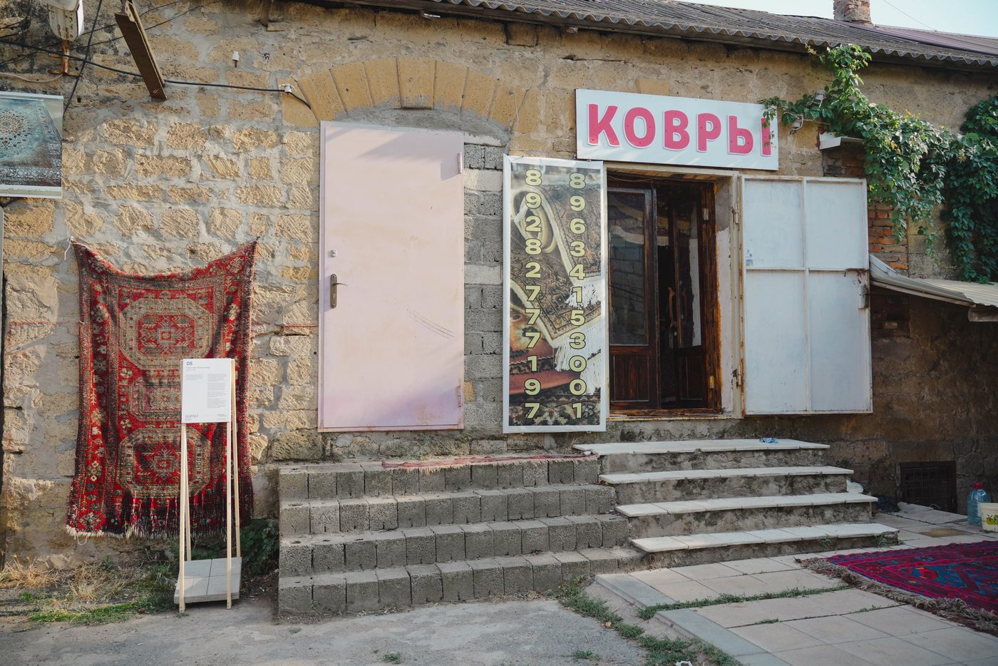 Фасад коврового магазина с вывеской «Ковры»: красный ковёр на стене, у входа — этикетка работы на деревянной стойке